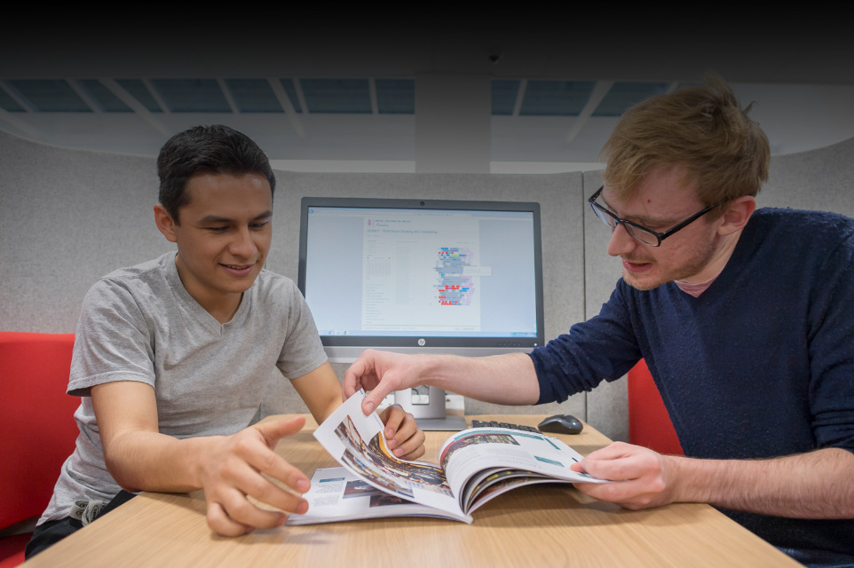 Two male students, one with glasses, looking through a prospectus, sitting in a group study pod.
